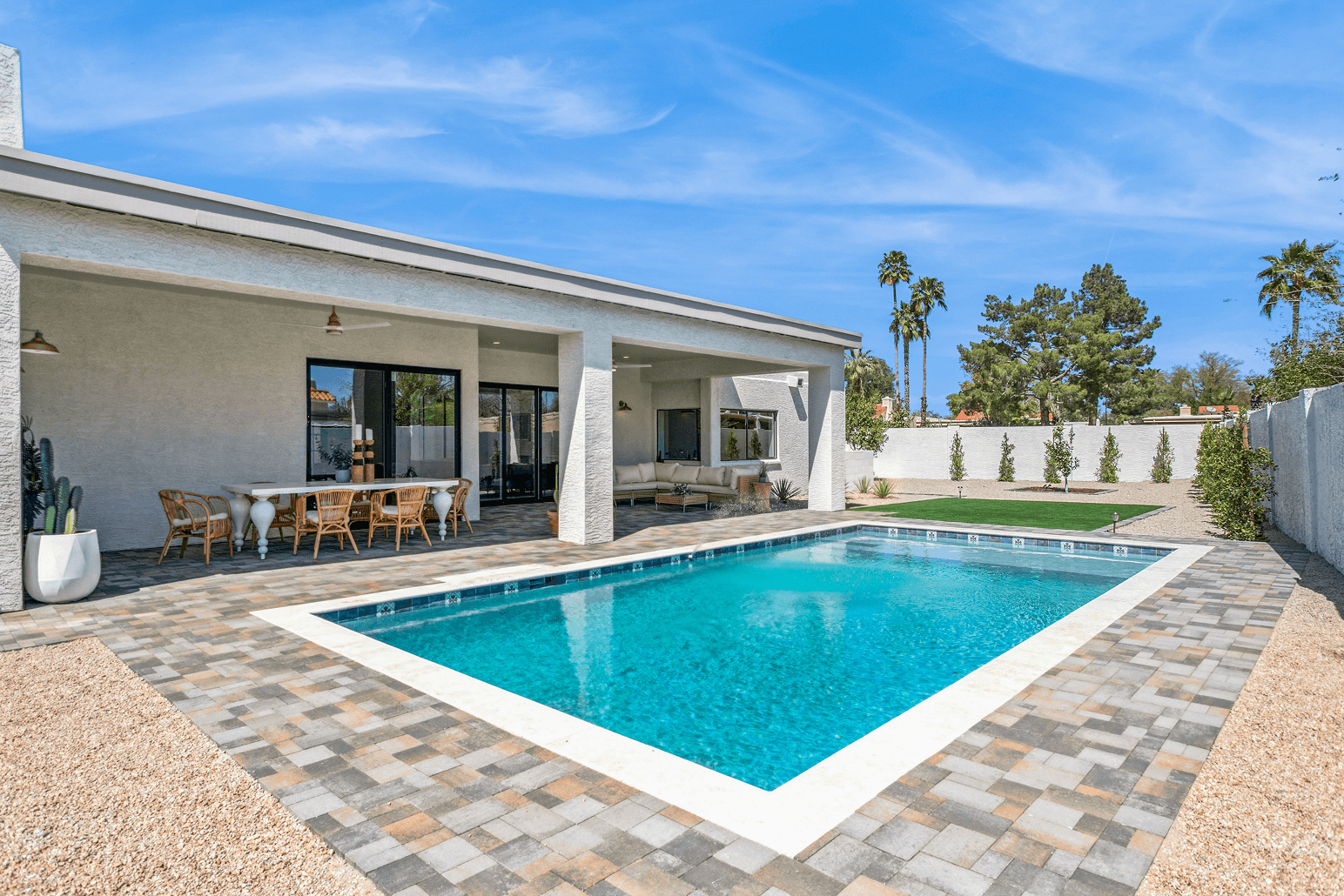 A modern backyard features a refreshing swimming pool, a covered patio with outdoor dining and seating, surrounded by pavers and desert landscaping under a blue sky.