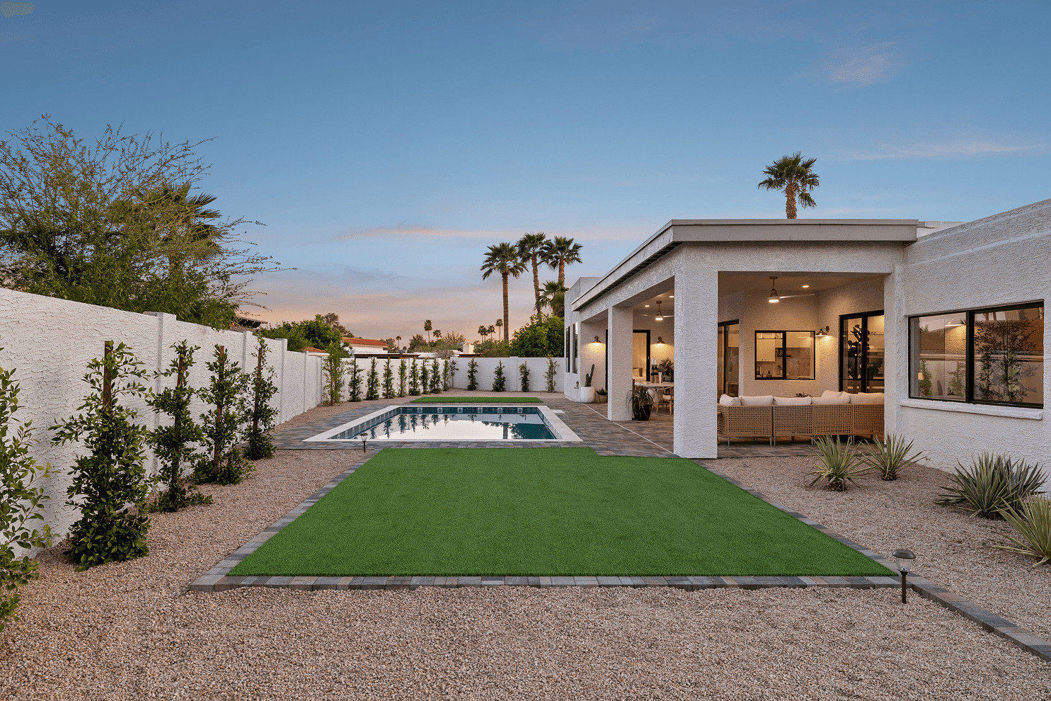 Backyard at dusk featuring a rectangular pool, artificial turf lawn, gravel landscaping, and a covered patio with outdoor seating and warm lighting.