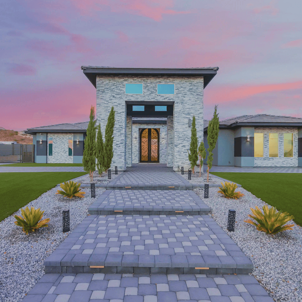 A modern house with a grey stone facade and a paver pathway leading to ornate double doors, set against a pink sunset sky.