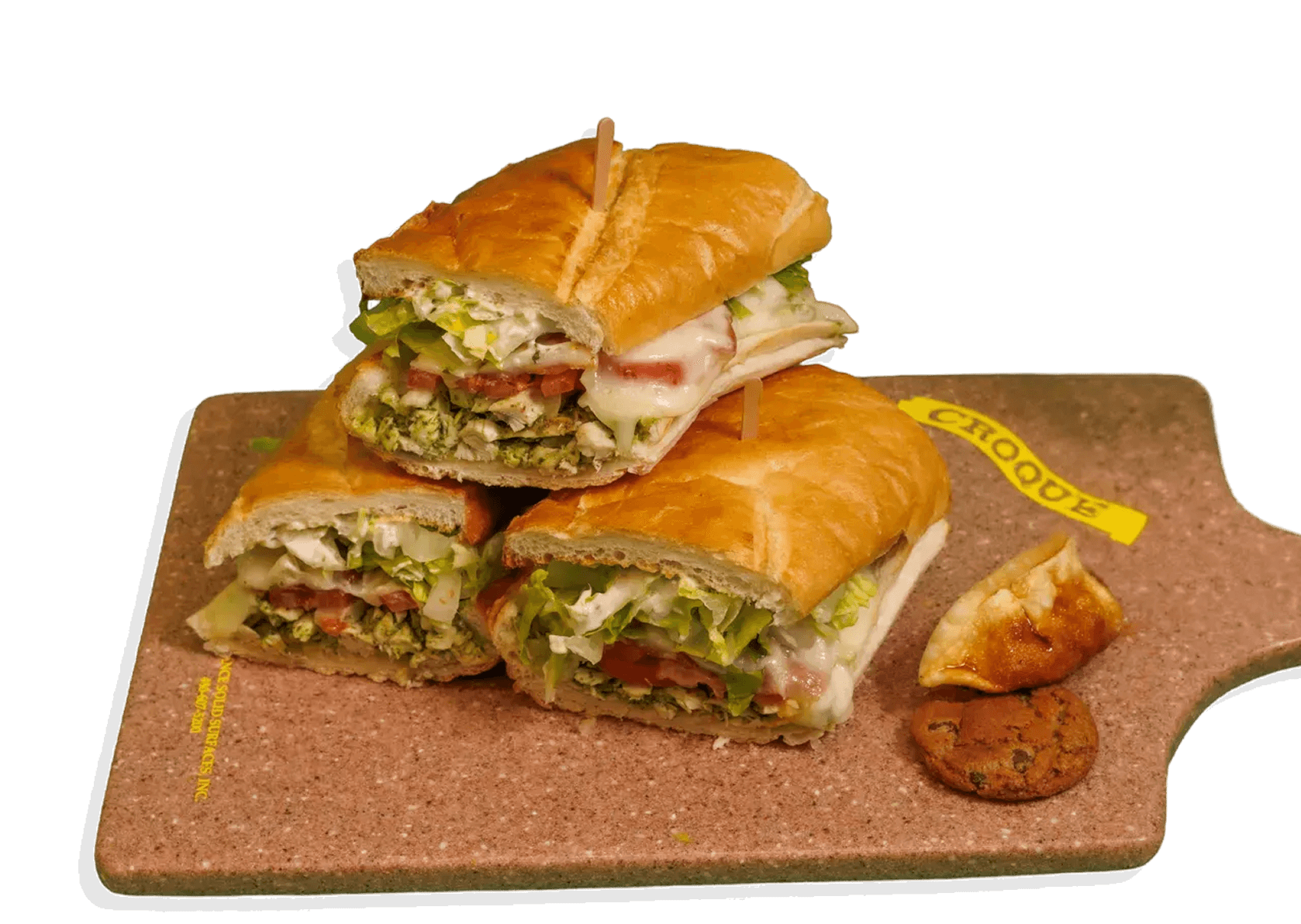 Three chicken pesto club sandwiches, cut in half, stacked on a brown cutting board with a cookie, a pastry, and a 'CROQUE' tag.