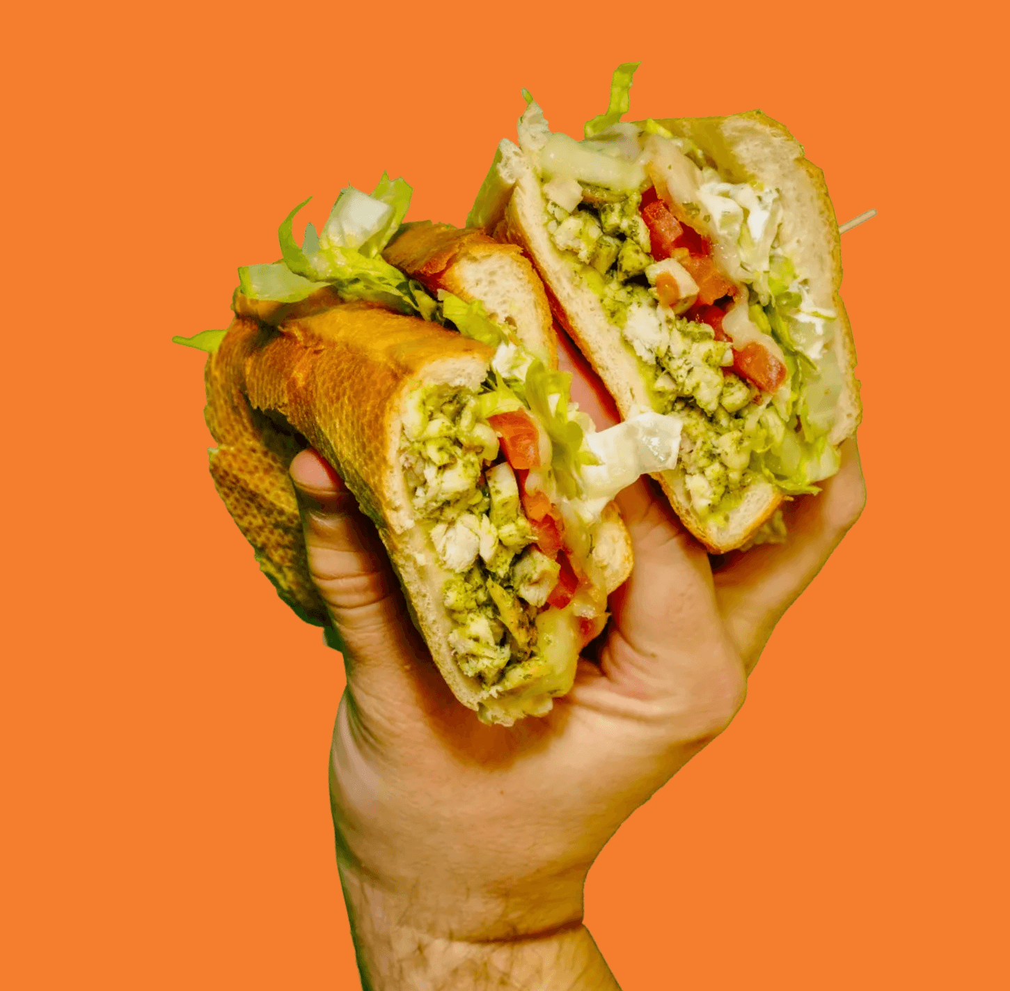 A hand holds a submarine sandwich cut in half, revealing chicken, pesto, lettuce, and diced tomatoes against an orange background.
