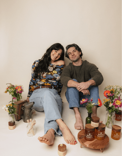 Smiling founders of 'Good Honey' sit on the floor with jars of honey, flowers, and beekeeping equipment, showcasing their natural product.