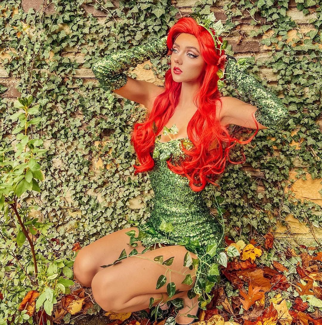 A woman cosplaying as Poison Ivy, with vibrant red hair, a sparkling green sequined dress, and ivy vines, crouches against an ivy-covered wall with autumn leaves.