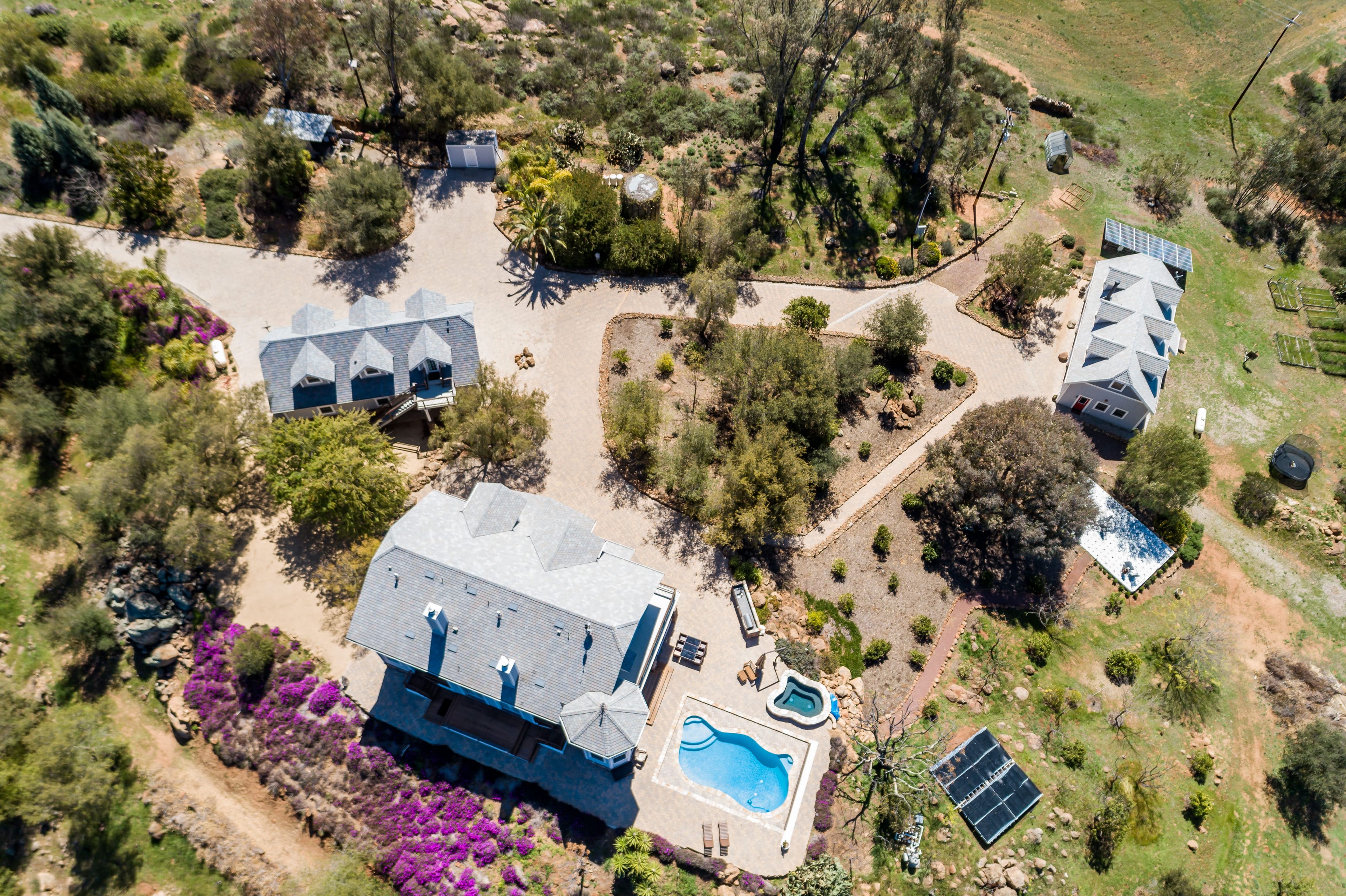 Aerial view of a multi-building residential property with a pool, hot tub, solar panels, and gardens, surrounded by natural rocky and vegetated terrain.