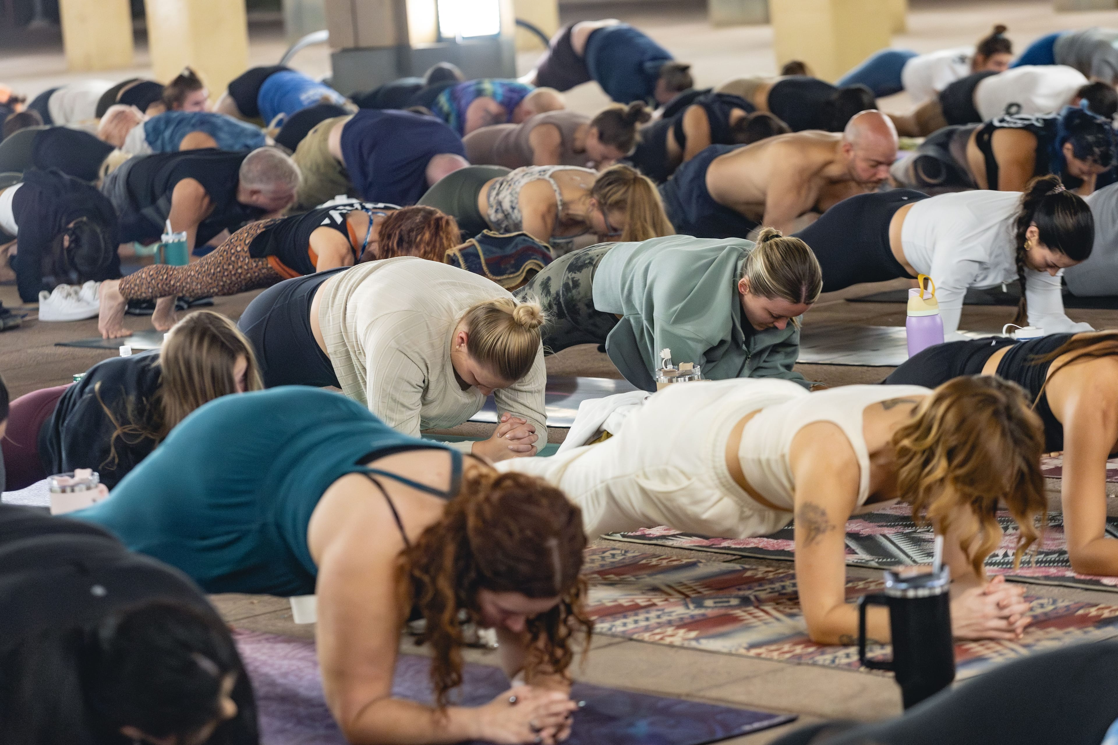 A large group of diverse people on yoga mats in an indoor space, supporting themselves on their forearms in a prone exercise pose.