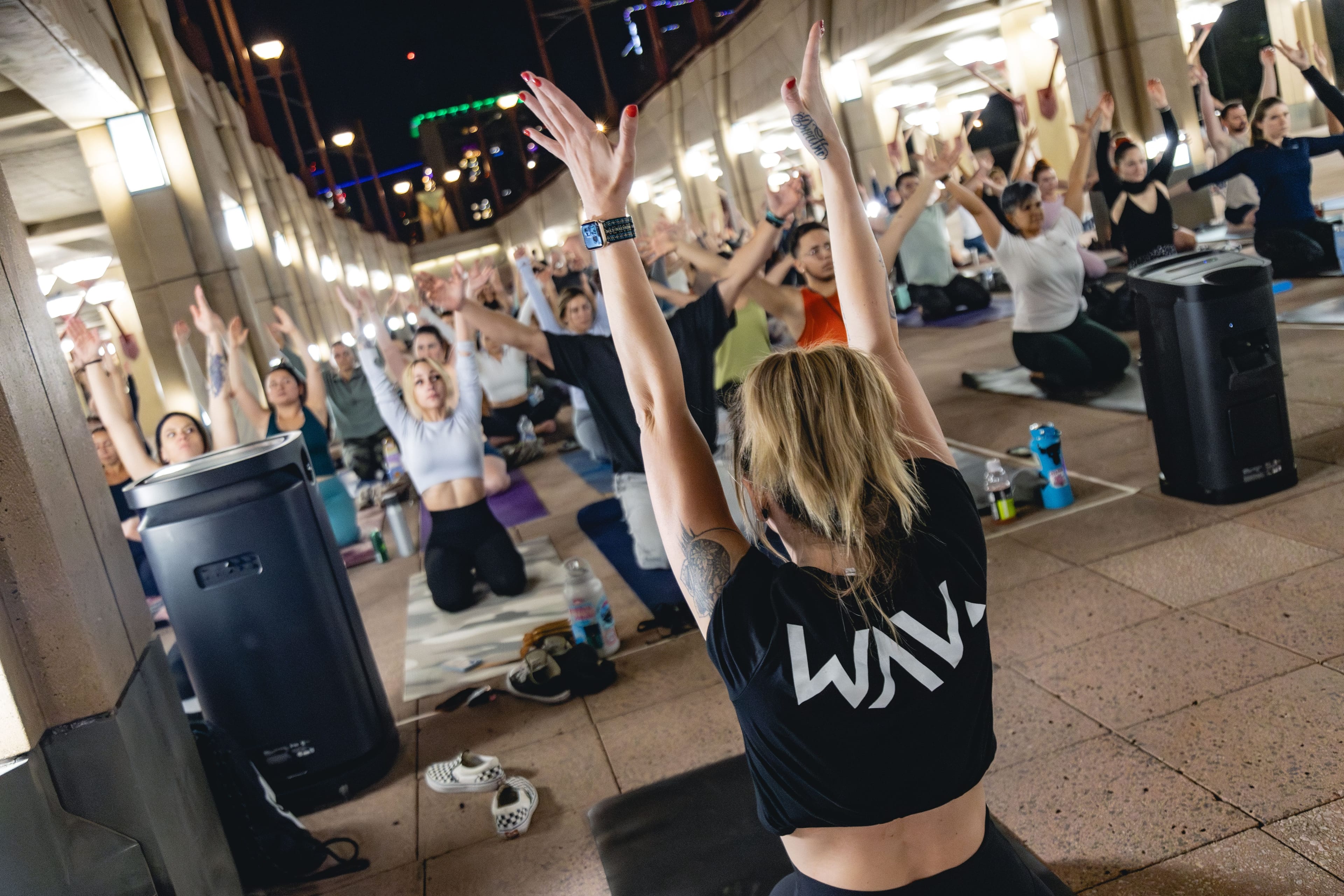 A diverse group of people participates in an evening outdoor yoga or fitness class under an illuminated concrete structure, raising their arms skyward. The foreground shows a participant from behind, wearing a black crop top with white text "WAVM".