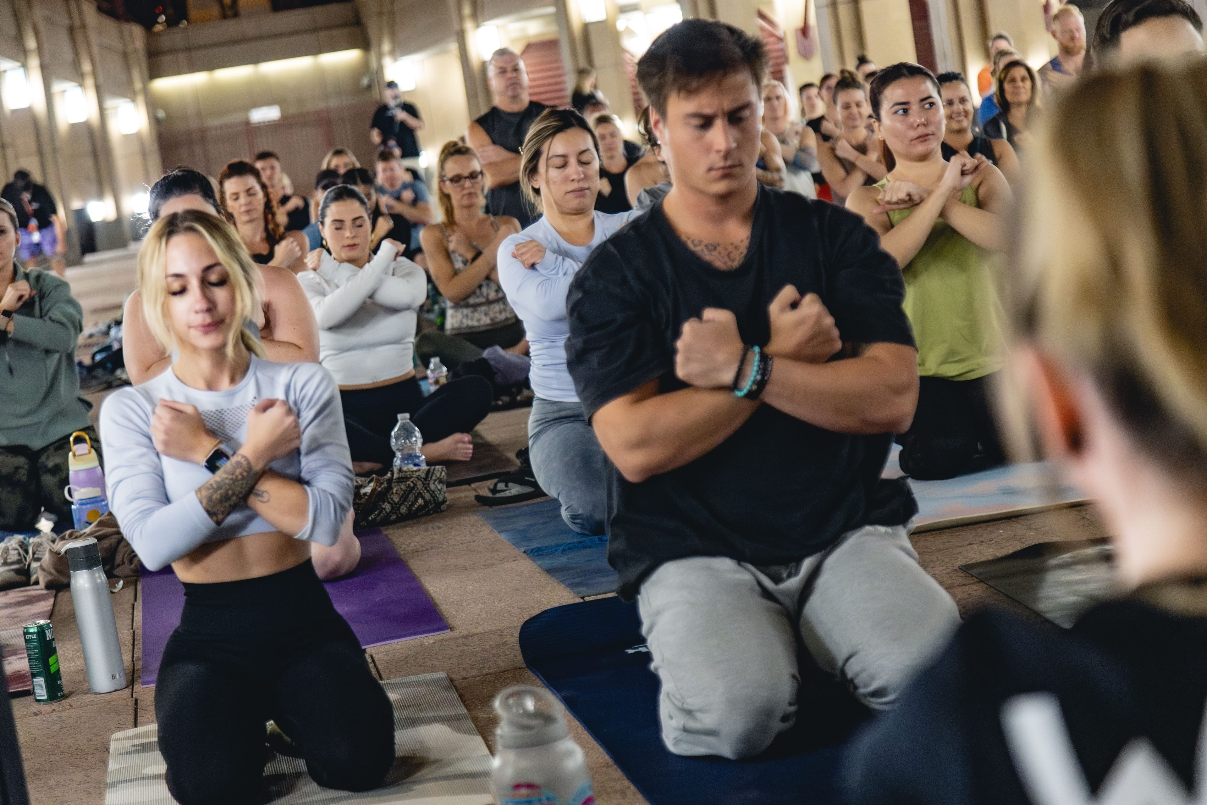 A group of people, primarily young adults, meditate or practice yoga indoors, kneeling on mats with their arms crossed over their chests in a focused pose.