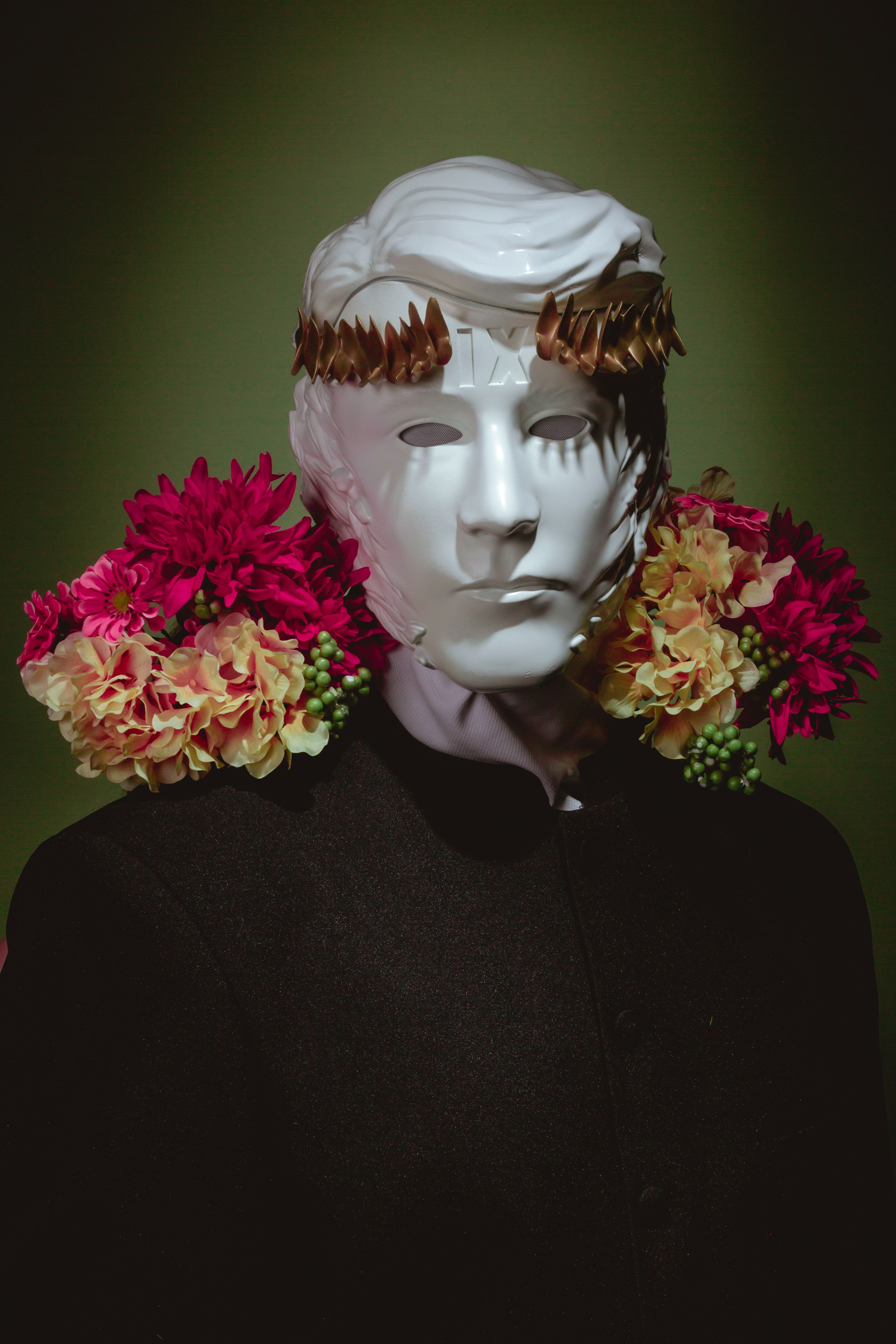 A white, stylized mask with a sculpted hairstyle resembling Donald Trump and a golden laurel crown, is draped with vibrant pink and yellow flowers, against a dark green background.