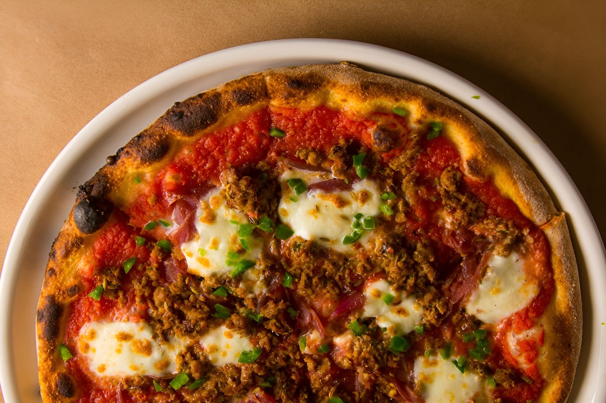 A close-up, top-down view of a well-baked pizza with vibrant tomato sauce, crumbled sausage, fresh mozzarella, and chopped green herbs, served on a white plate.