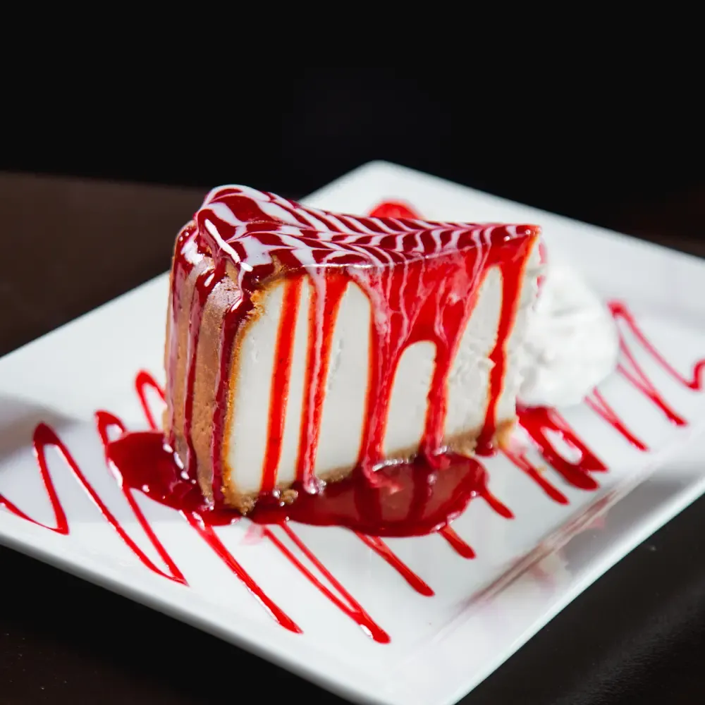 A slice of cheesecake drizzled generously with bright red fruit sauce and a dollop of whipped cream, presented on a white rectangular plate.