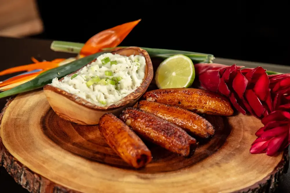 Candied plantains and a bowl of creamy dip with green onions, garnished with lime and tropical flowers on a wooden platter.