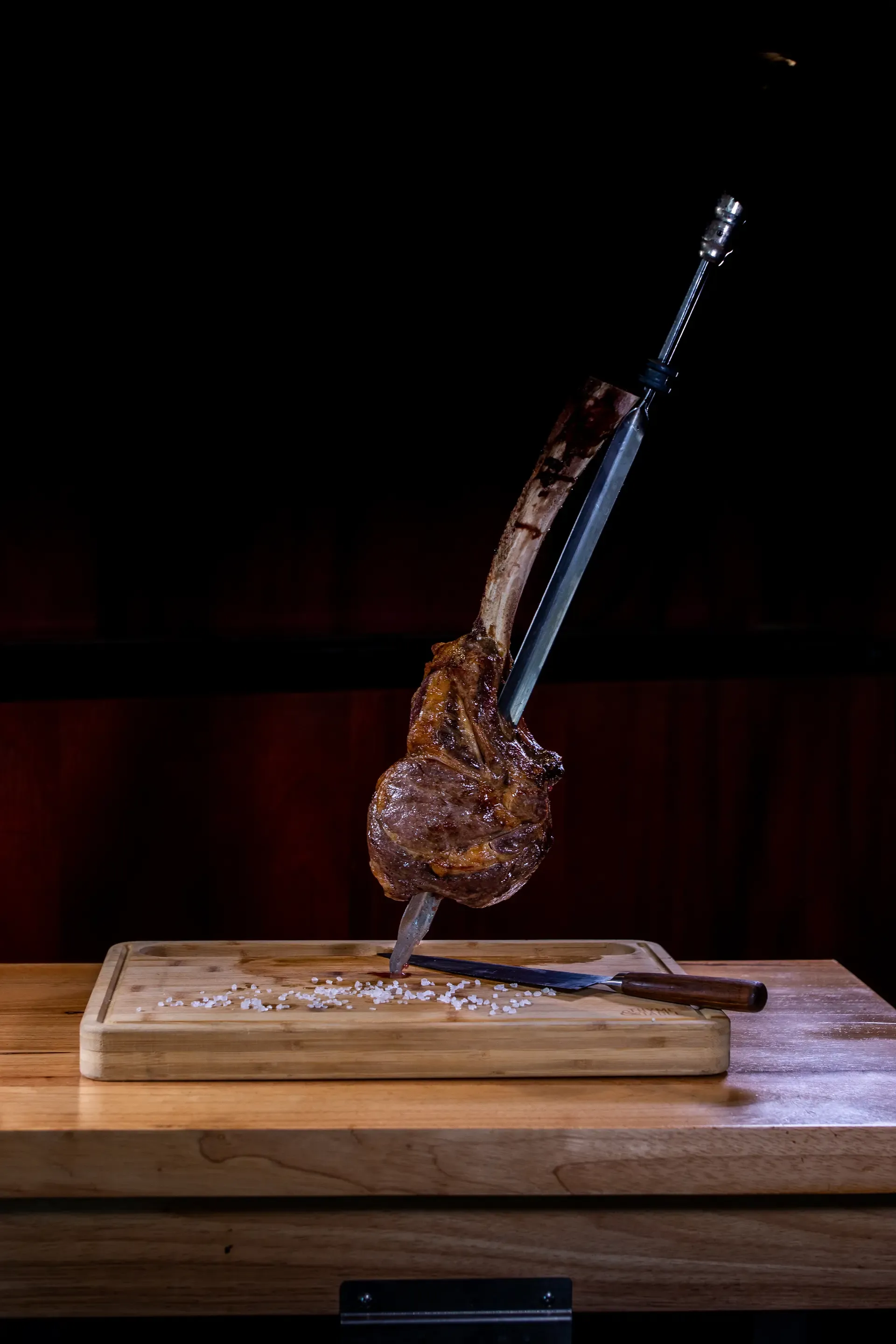 A large, perfectly cooked tomahawk steak with the bone stands vertically, impaled on a metal skewer on a wooden cutting board, surrounded by coarse salt and a knife.