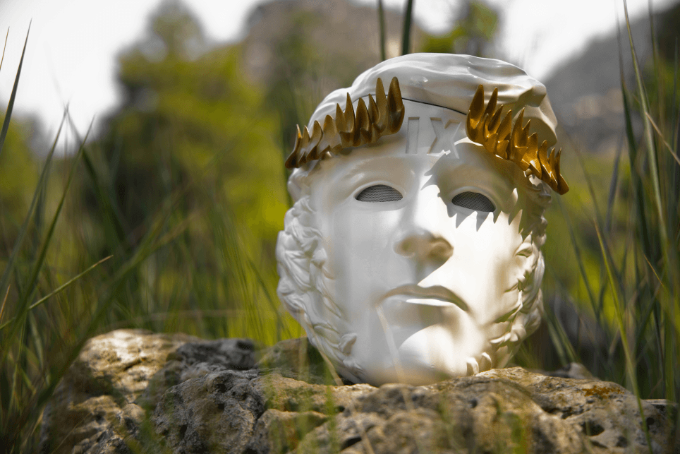 A white classical-style mask with a gold spiky crown and "IX" on its forehead, resting on a sunlit rock amidst tall green grass.