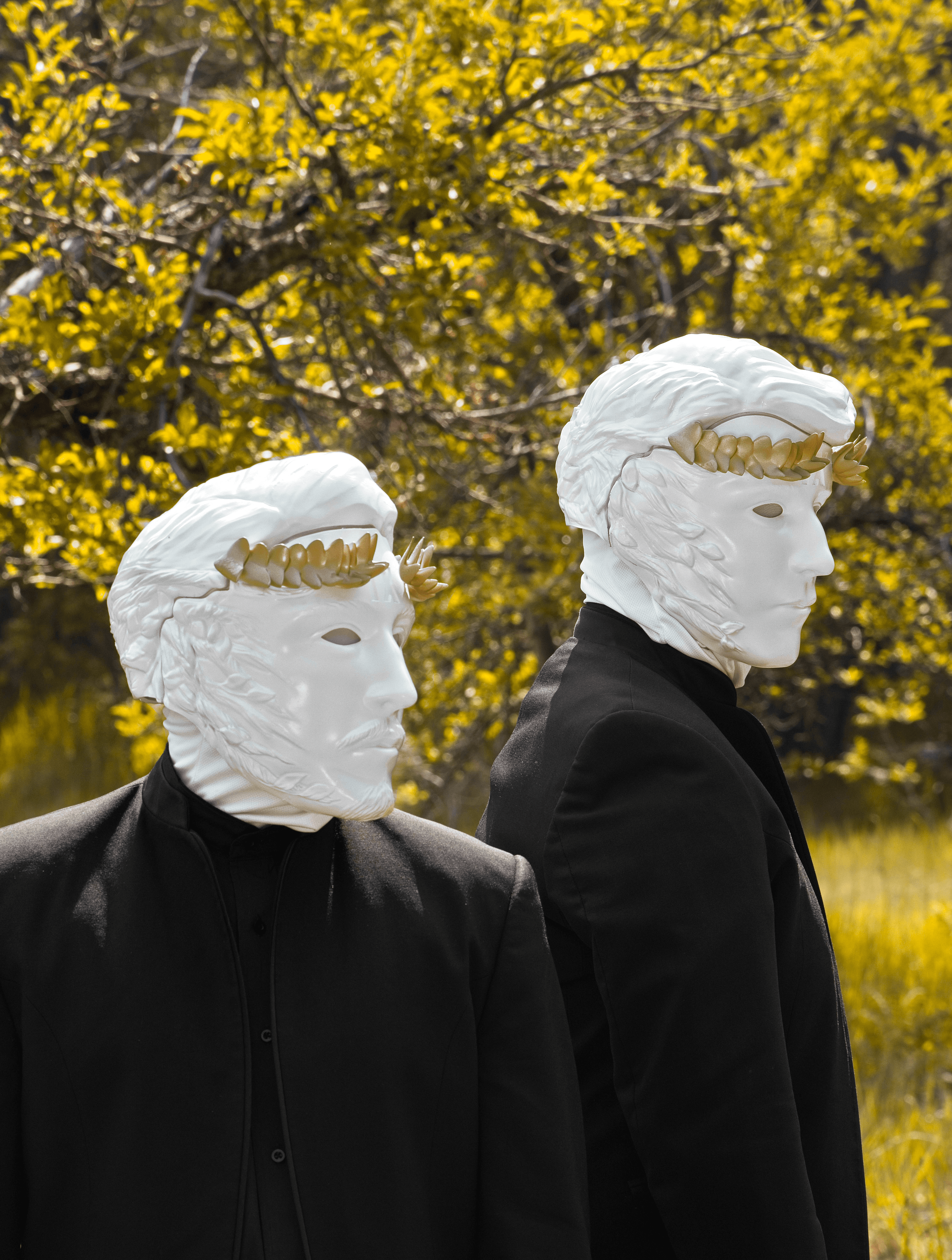 Two figures in black wear white, sculpted masks resembling classical busts with golden laurel wreaths, facing opposite directions in a blurry yellow-green landscape.