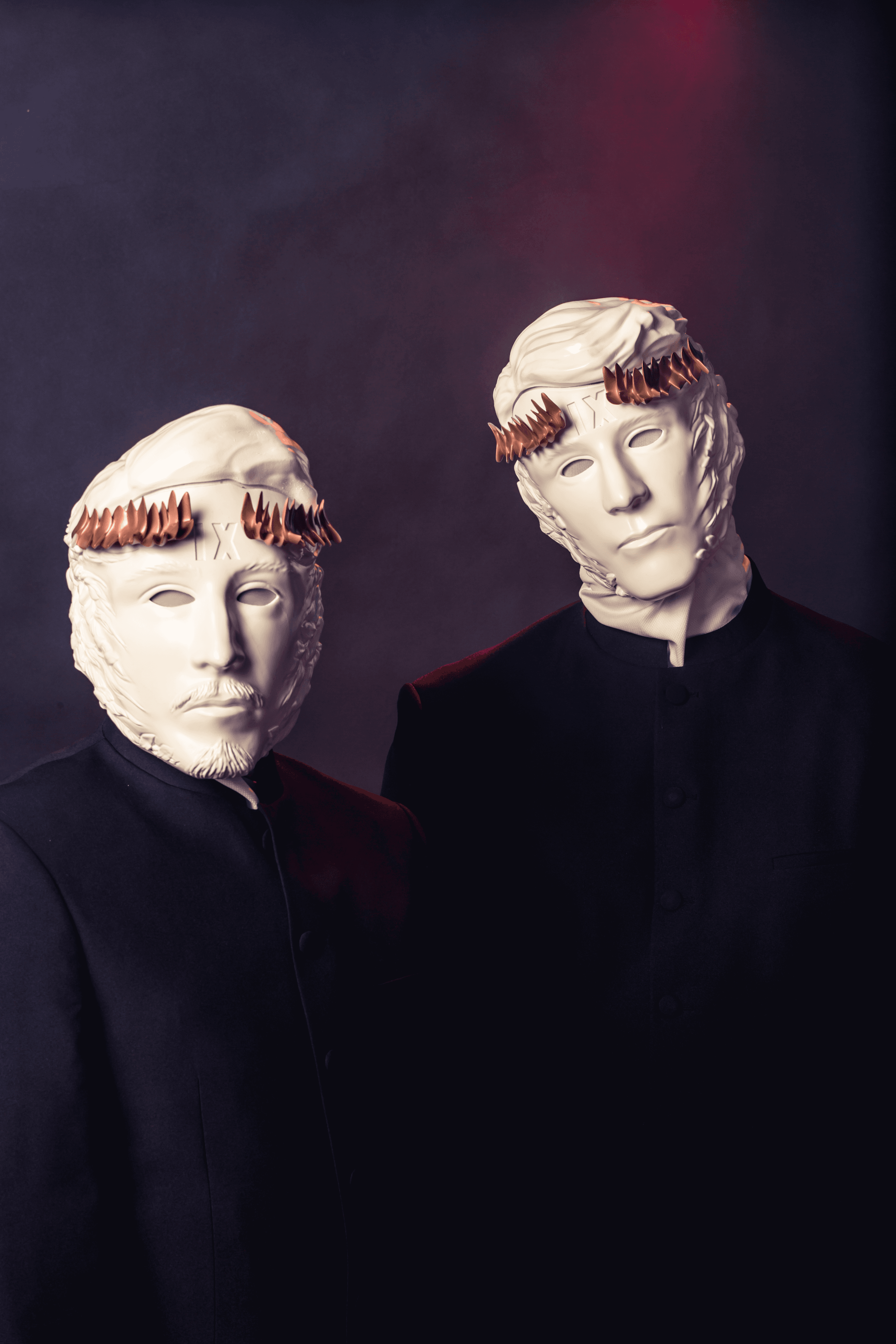 Two figures in dark priest-like suits wear white, sculpted masks; one has a beard, the other "IX" and both feature spiked bronze crowns, against a dark, red-lit background.