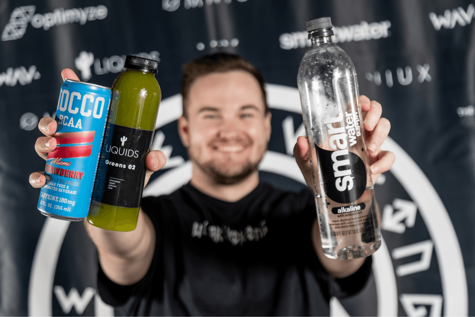 A smiling man holds up a blue Roccoco BCAA can, a green LIQUIDS Greens 02 bottle, and a smartwater alkaline bottle in front of a branded backdrop.