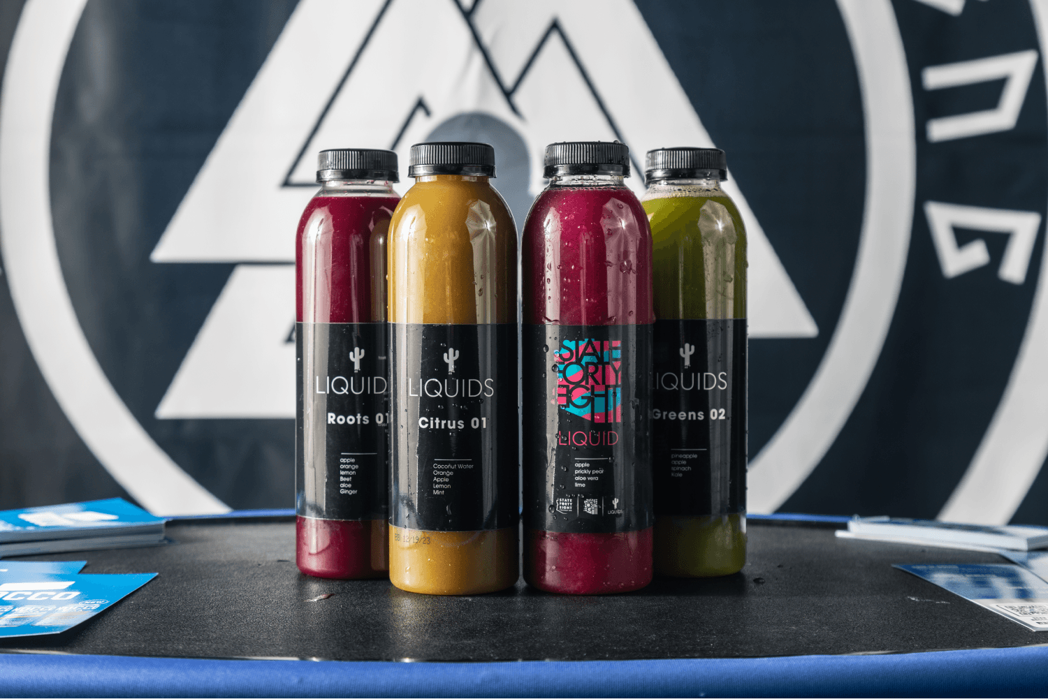 Four bottles of colorful cold-pressed juices from "Liquids" and "State Forty Eight" brands, displayed on a dark table in front of a geometric logo banner.