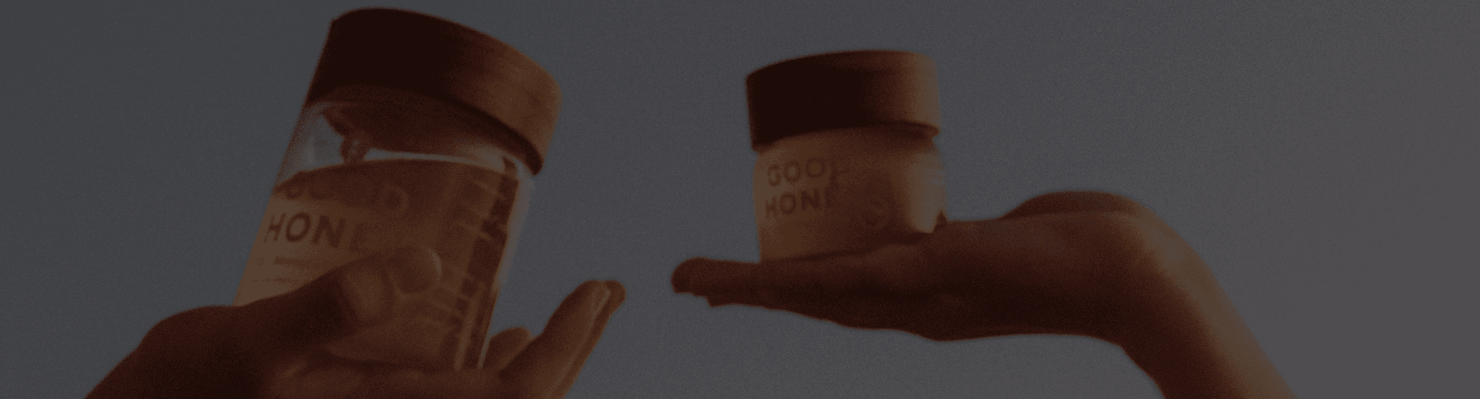 Two hands hold two jars of honey, glowing warmly in dim light, with "GOOD HONEY" visible on their labels and wooden caps, against a dark background.