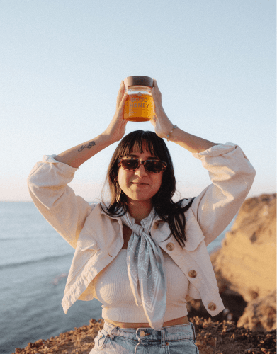 A smiling woman in sunglasses and a jacket holds a jar labeled "GOOD HONEY" above her head on a sunlit cliff overlooking the ocean.
